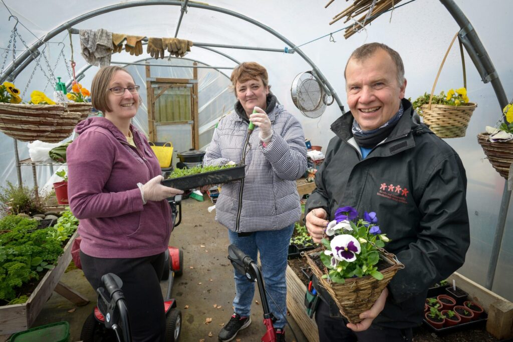 3 smiling people in a greenhouse holding flowers and seedlings.
