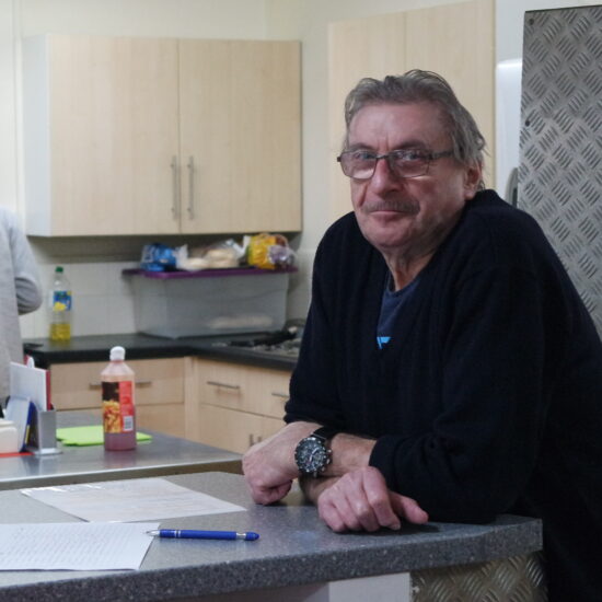 Man sitting at table in a community kitchen, woman at sink in background