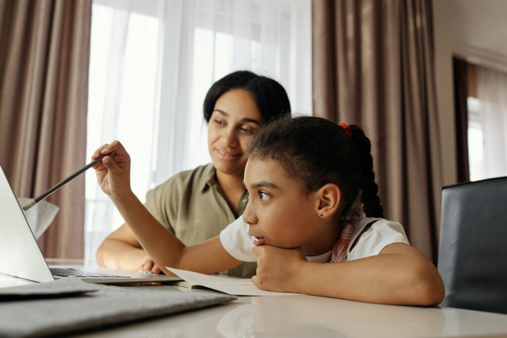Mother and daughter talking to someone via a laptop. The little girl is holding a pencil and talking.