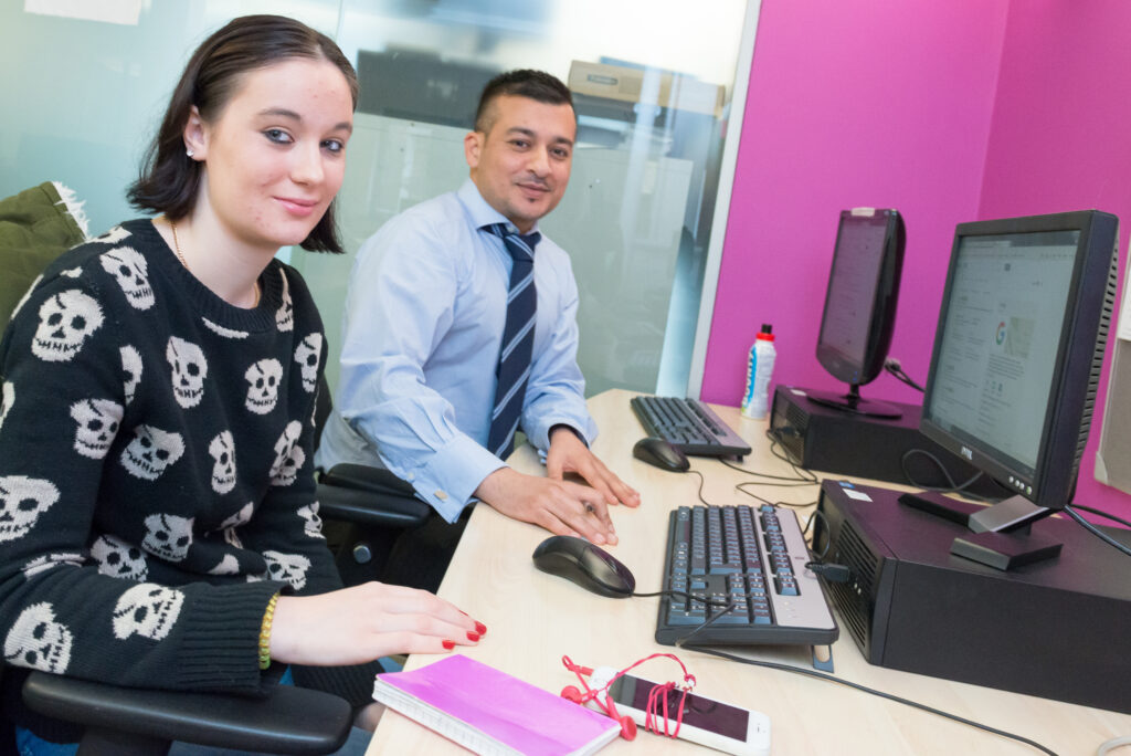 Two people sitting together at a computer, smiling towards the camera.