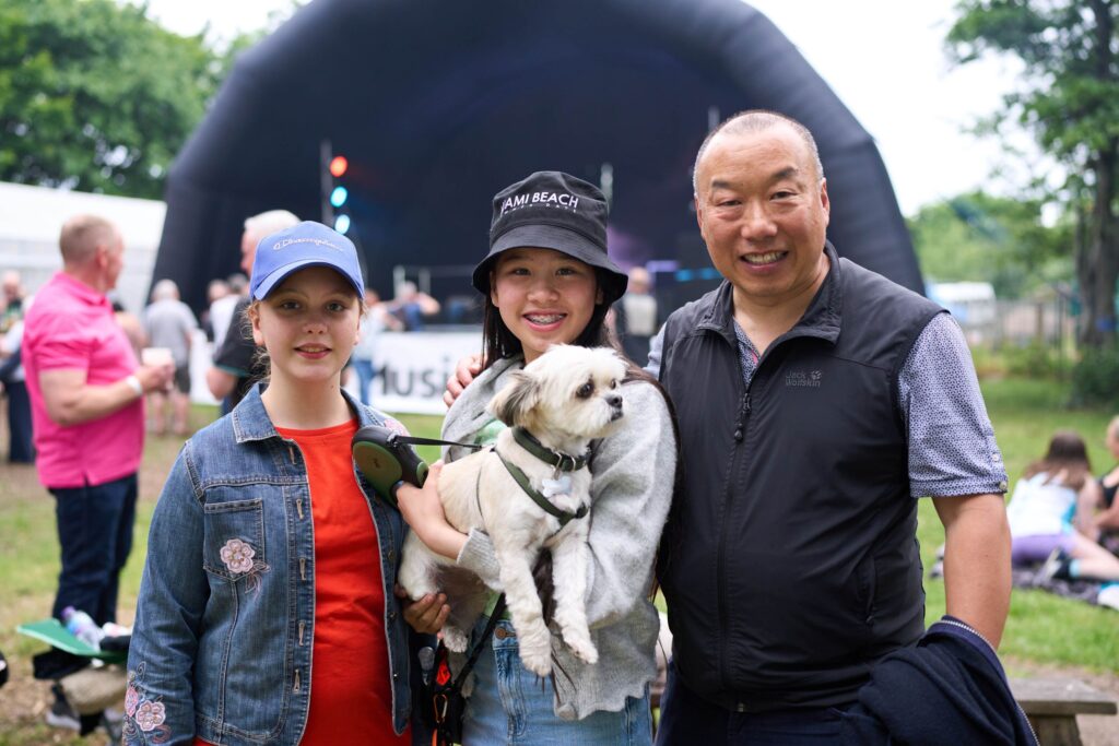 Two young people, wearing hats one holding a white dog, and a man. They're all standing in front of an outdoor stage,