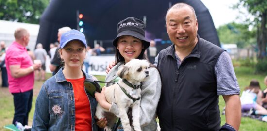 Two young people, wearing hats one holding a white dog, and a man. They're all standing in front of an outdoor stage,