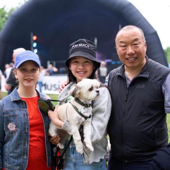 Two young people, wearing hats one holding a white dog, and a man. They're all standing in front of an outdoor stage,