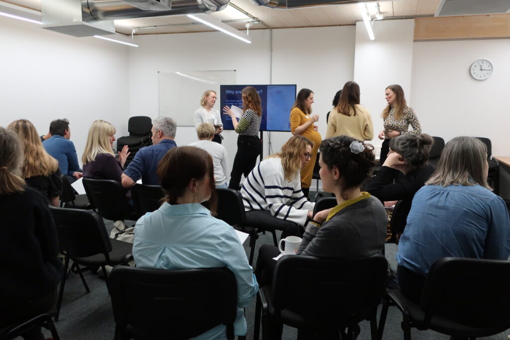 Community of practice in person meeting. Lots of people seated looking towards the front of the room where Renaisi's staff are standing in front of a TV screen.
