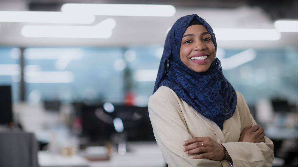 A woman wearing a hijab, cross-armed and smiling in an office environment.